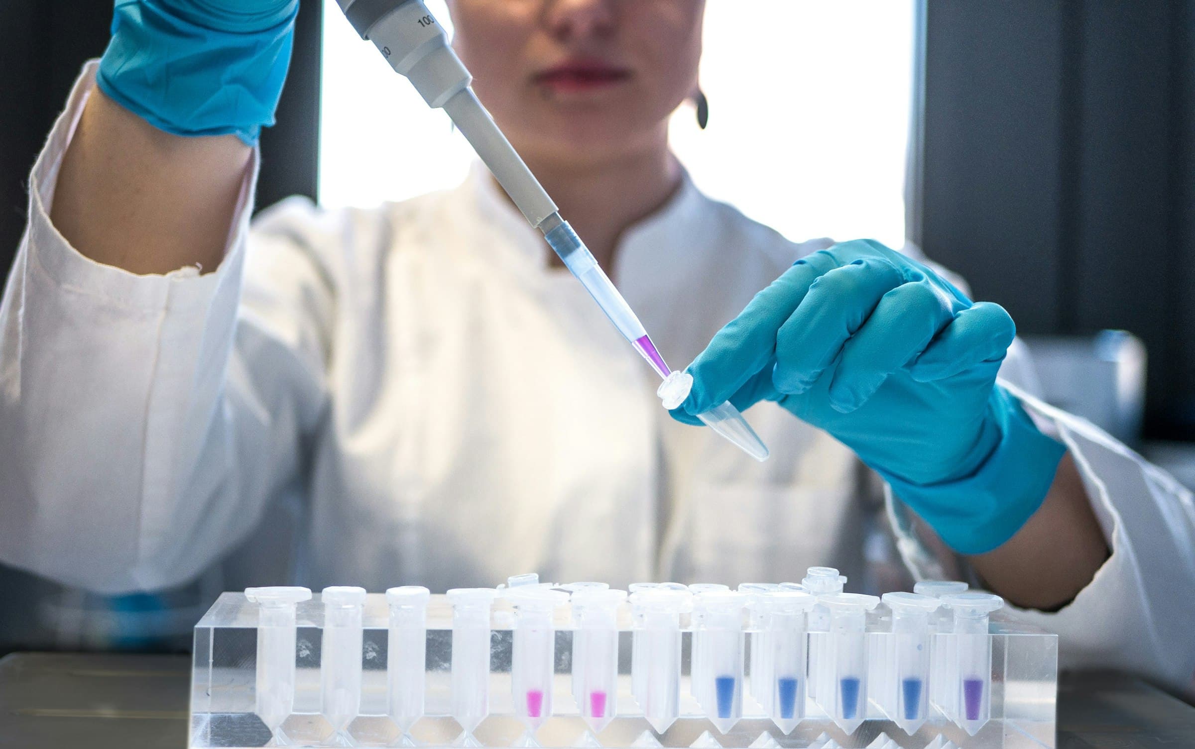 Pesquisadora de jaleco branco e luvas azuis utilizando pipeta multicanal sobre um suporte de microtubos em laboratório